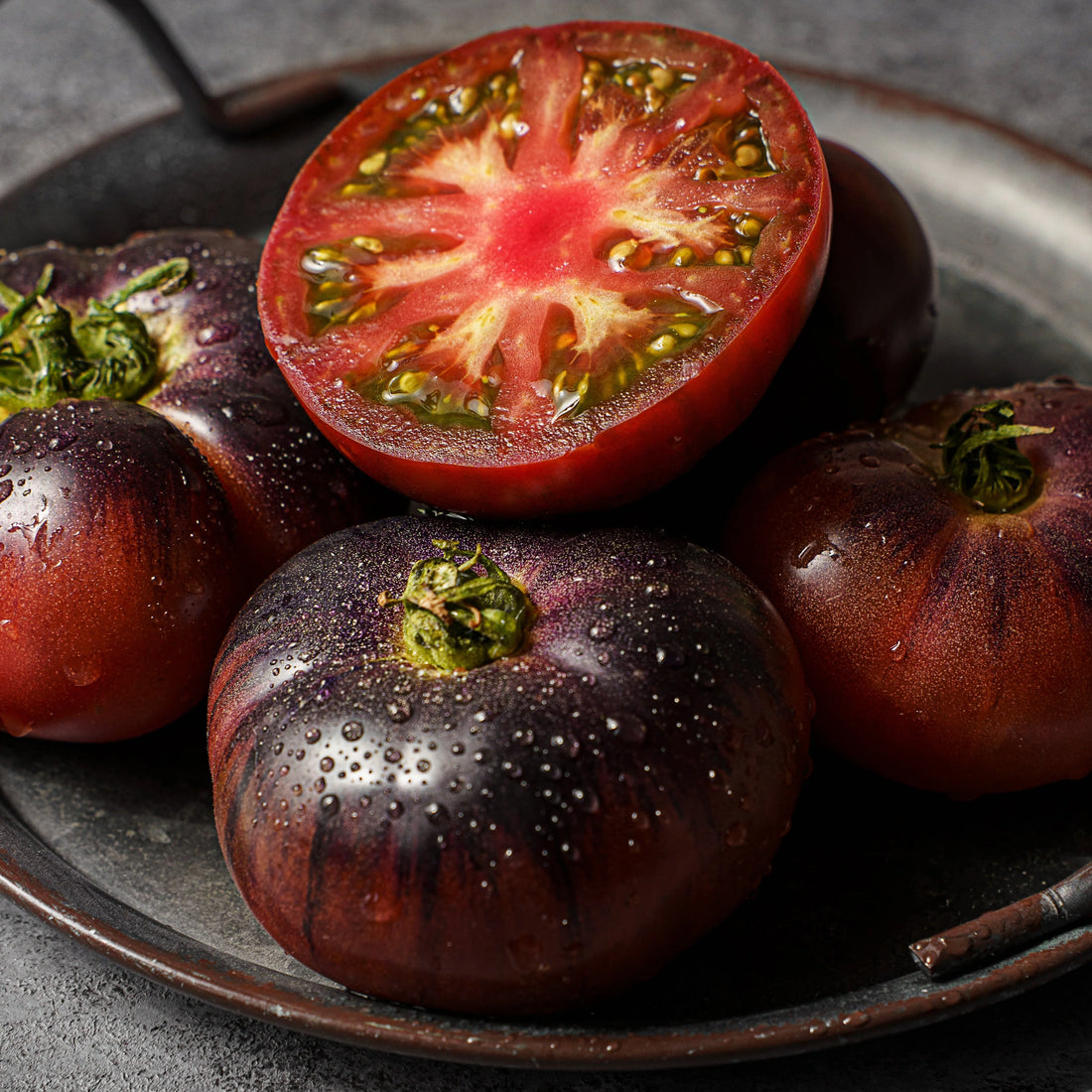 Black tomatoes on a dark plate with one sliced open to reveal red flesh.